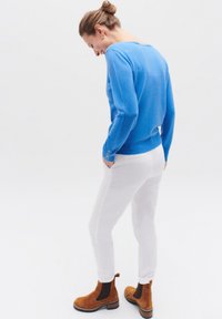 Blauwe gebreide trui met lange mouwen, witte strakke broek en bruine suède enkelboots. Opvallende knoopdetails op de mouw. Eenvoudig ontwerp.