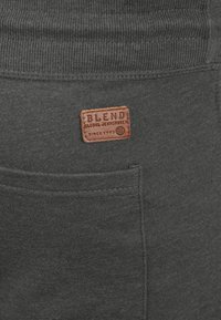 Grå stofbukser med ribbet talje og en brun læderlap med teksten "BLEND GLOBAL JEANMAKER" syet på den bageste lomme.