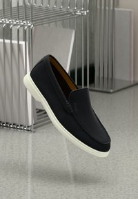 Sorte slip-on loafers med hvit gummisåle, vist på et reflekterende gulv nær et metalltrådstativ.
