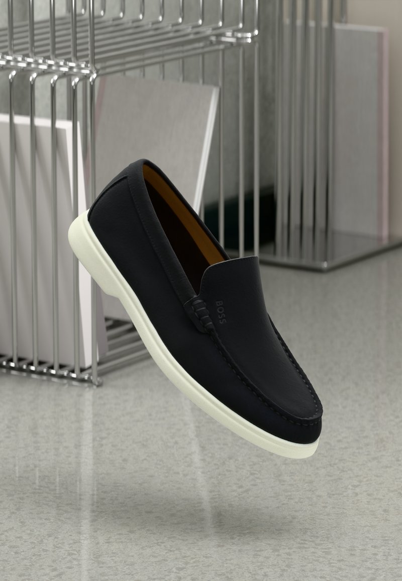 Sorte slip-on loafers med hvit gummisåle, vist på et reflekterende gulv nær et metalltrådstativ.