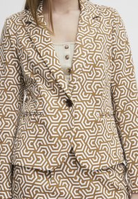 Brauner Blazer mit geometrischem Muster und weißen Akzenten, der eine taillierte Passform, Reverskragen und zwei Fronttaschen bietet. Knöpfe sind am Kragen sichtbar.