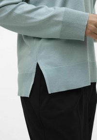 Maglione azzurro chiaro con polsini e orlo a coste, dotato di spacchi laterali. Abbinato a pantaloni neri, che mostrano una texture a maglia liscia.