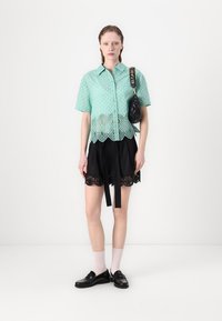 Mintgroene geperforeerde blouse met een gegolfde zoom, gecombineerd met zwarte shorts met kantafwerking en strikjes. Zwarte schoenen maken de outfit compleet.