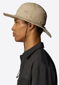 Sombrero de cubo beige con ala ancha, hecho de material ligero, presenta una correa ajustable para el mentón y un logo bordado en la parte frontal.