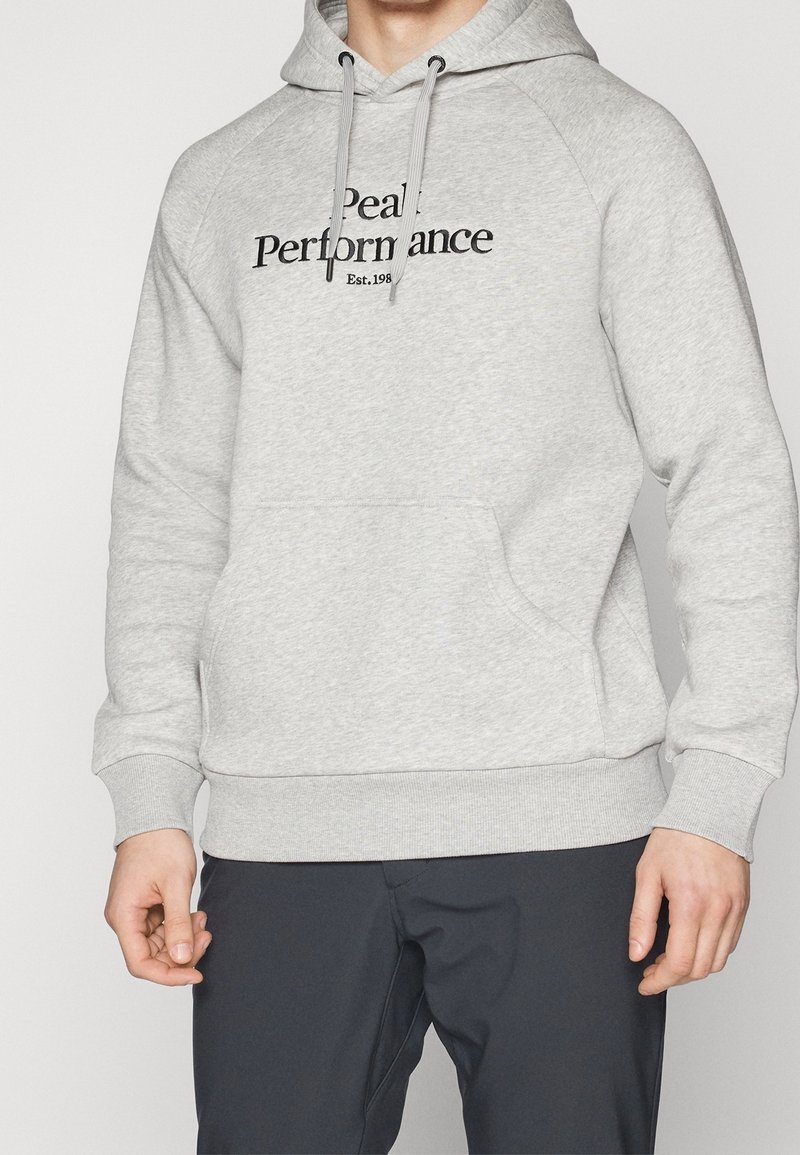 Hoodie gris clair avec une poche kangourou, des poignets côtelés et un bas en bord côte ; arbore le logo "Peak Performance" en noir. Texture de tissu douce.