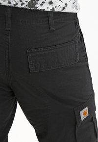 Pantalons cargo noirs en tissu texturé, avec une poche arrière et une étiquette de logo Carhartt orange et blanche sur le côté.