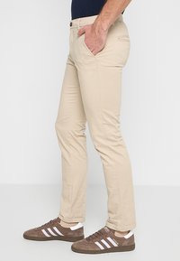 Bărbat îmbrăcat în pantaloni slim-fit bej și adidași maro de la Adidas, stând cu o mână în buzunar pe fond alb.
