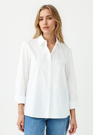 Femme blonde portant une chemise blanche à boutons avec les manches retroussées et un jean bleu, se tenant contre un fond blanc uni.
