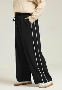 Svarta vida byxor med vita sidskarvar, med resårmidja och dragsko. Matchas med vita sneakers på en neutral bakgrund.