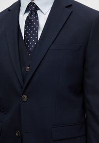 Costume bleu marine en tissu texturé, design à une seule boutonnière, et une poche. Associé à une cravate bleue marine à pois roses.