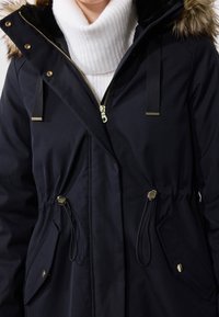 Czarny zimowy płaszcz z złotym zamkiem błyskawicznym i zatrzaskami, regulowanym sznurkiem w talii, kieszeniami z patkami oraz kapturem obszytym futrem, noszony na białym swetrze z golfem.