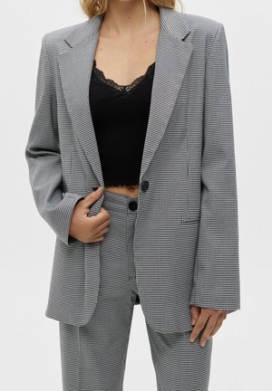 Vrouw draagt een zwart crop top met kanten rand onder een grijs-witte pied-de-poule blazer en bijpassende hoge taille broek, hand houdt het revers van de blazer vast.