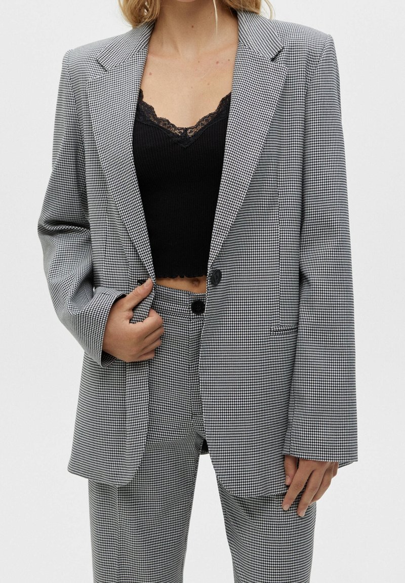 Femme portant un crop top noir bordé de dentelle sous un blazer pied-de-poule gris et blanc et un pantalon taille haute assorti, main tenant le revers du blazer.