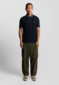 Polo shirt navy con colletto a fantasia e un piccolo logo giallo, abbinato a pantaloni cargo oliva e sneakers beige con accenti grigi.