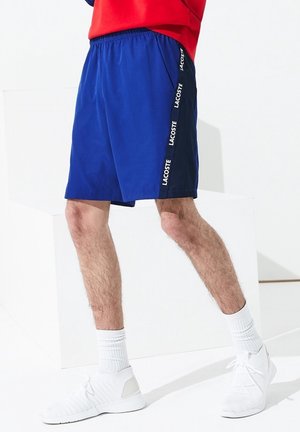 Mandlige ben iført blå Lacoste shorts, hvide sokker og hvide sportssko, stående mod en hvid baggrund.