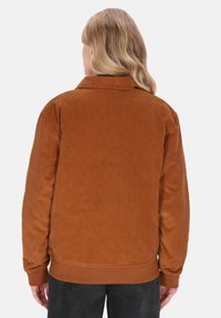 Persoon met lang blond haar, met de rug naar ons toe, gekleed in een bruine ribfluwelen jas en zwarte jeans, tegen een effen licht achtergrond.