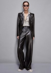 Completo in pelle nera con giacca oversize e pantaloni a gamba larga. Camicia color grigio chiaro con colletto sotto. Gli accessori includono occhiali da sole e orecchini a cerchio.