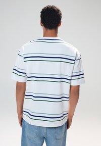 T-shirt bianca a maniche corte con righe orizzontali in blu scuro, azzurro e verde. Tessuto morbido con una vestibilità comoda, cuciture visibili.