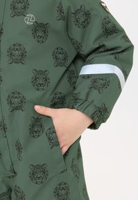 Groene regenjas met een patroon van zwarte geometrische leeuwenkoppen. Elastische manchetten, zijzak en reflecterende streep op de mouw.