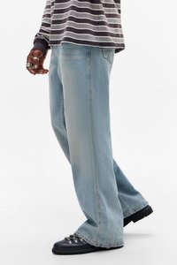 Person som bruker lyseblå vide jeans, svarte tursko og en langermet stripete skjorte, står mot en hvit bakgrunn.