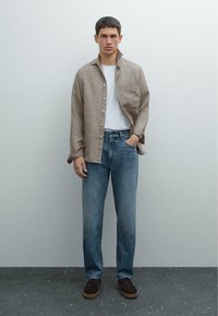 Bruin geruit overhemd, witte t-shirt eronder, blauwe spijkerbroek en donkerbruine suède schoenen. Model staat tegen een neutrale achtergrond.