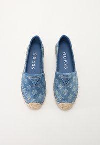 Denim espadrilles met blauwe patroonstof, een driehoekig metalen logo-accent en een natuurlijke geweven jute zool. Platter ontwerp, instapstijl.