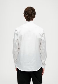 Man med lockigt brunt hår som bär en långärmad vit skjorta och svarta byxor, sedd bakifrån mot en enfärgad bakgrund.