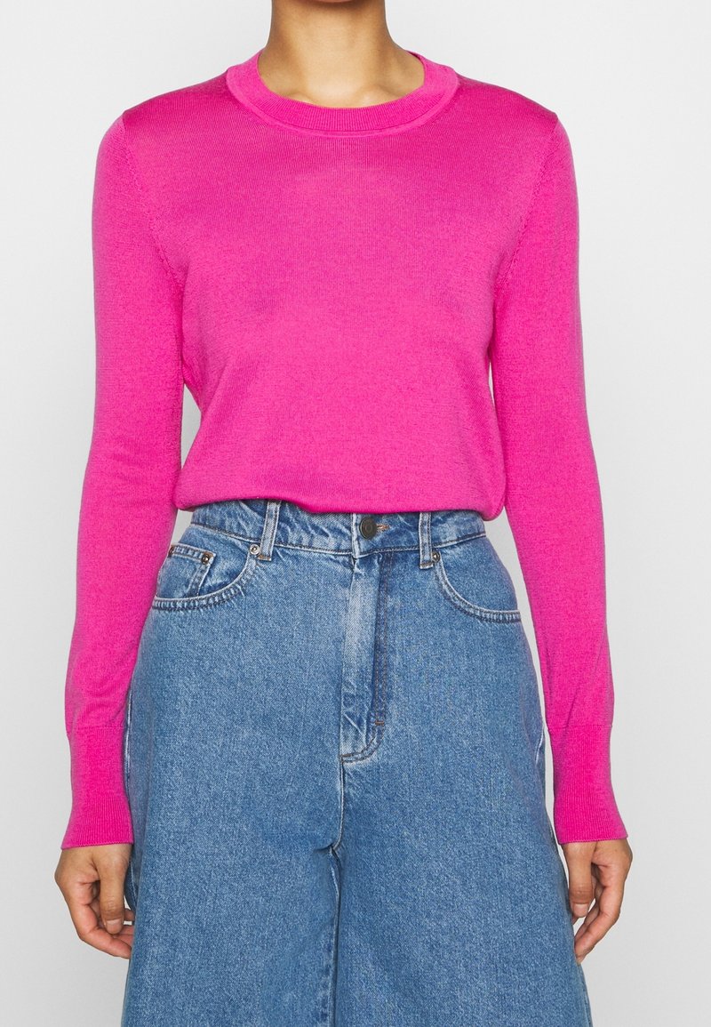 Pull tricot à manches longues fuchsia avec un col rond, présentant une texture lisse. Associé à un jean denim bleu taille haute à jambes larges.