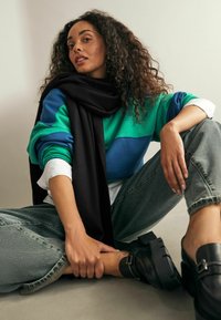 Sweatshirt coloré avec des rayures vertes et bleues sur une chemise blanche, associé à un jean taille haute et des chaussures compensées noires. Écharpe noire drapée.