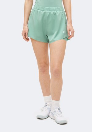 Γυναίκα που φοράει ανοιχτά πράσινα αθλητικά σορτς, λευκές κάλτσες μέχρι τον αστράγαλο και λευκά αθλητικά παπούτσια, στέκεται σε απλό ανοιχτό φόντο.