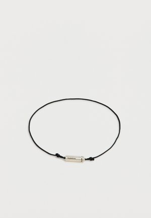 Schwarzes verstellbares Kord-Armband mit einem kleinen zylindrischen silbernen Verschluss, graviert mit „le gramme“ auf weißem Hintergrund.