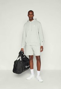 Man wearing light gray hoodie and shorts holding black duffel bag, white socks, and white sneakers, standing against plain background.