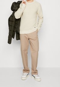 Ljusbeige stickad tröja med rund halsringning, kombinerad med ljusbruna byxor. En mörkgrön pufferjacka hänger över ena armen. Vita sneakers.