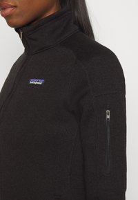 Zwarte zip-up jack met een hoge kraag, gebreide textuur, zijzak met rits en een Patagonia-logo in blauw en paars vlakbij de borst.