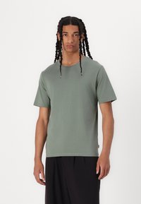 Tricou verde cu mânecă scurtă, realizat dintr-un material moale, cu o croială lejeră, având un guler rotund și detalii de cusătură subtile.