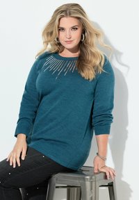 Tealfarbener Pullover mit langen Ärmeln und Rundhalsausschnitt, der metallische Akzente in einem Sonnenstrahlenmuster auf der Brust aufweist; kombiniert mit dunklen Jeans.