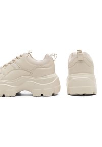 Beżowe sneakersy z grubą, masywną podeszwą, teksturowaną cholewką i okrągłym noskiem. Oferują minimalistyczne sznurowanie i tylną pętelkę ułatwiającą zakładanie.