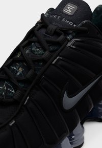 Κοντινή λήψη μαύρου αθλητικού παπουτσιού Nike Shox που δείχνει τα κορδόνια, το γκρι σήμα Nike, υφασμάτινη υφή και λεπτομέρειες της μαξιλαρωμένης σόλας.