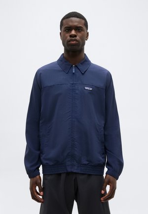 Mees, kes kannab tumesinist lukuga jope, millel on krae ja elastne ääreosa, seisab tavalise heleda tausta ees.