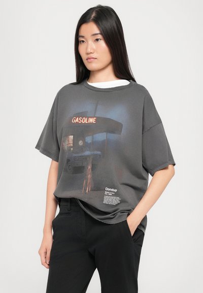 T-shirt oversize gris avec un imprimé graphique d'une station-service, comportant le texte rouge "GASOLINE" et une étiquette avec des détails de marque sur l'ourlet.