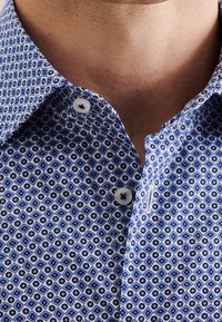 Mužský krk a horní část hrudníku oblečený v košili s modrým vzorem a bílými knoflíky, límec otevřený, ukazující kůži pod ním.