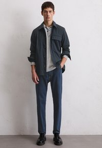 Marinejacke mit Brusttasche über einem hellgrauen Poloshirt, kombiniert mit dunkelblauer Jeans und schwarzen Schuhen, vor einem neutralen Hintergrund.