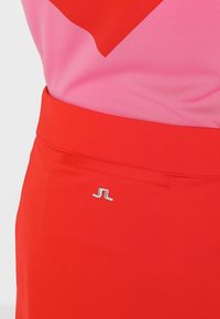 Červená sukně s hladkou texturou a přiléhavým pasem. Má malý stříbrný logotyp poblíž pasové linie. Růžový top je částečně viditelný.