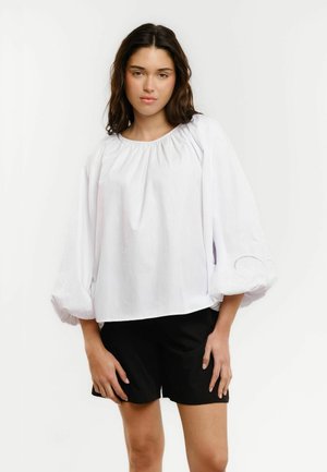 Woman wearing a white blouse with balloon sleeves and black shorts, standing against a plain white background.