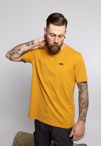 T-shirt a maniche corte giallo senape con tasca, scollatura rotonda e dettagli cuciti. Il modello ha tatuaggi ed è posizionato su uno sfondo neutro.