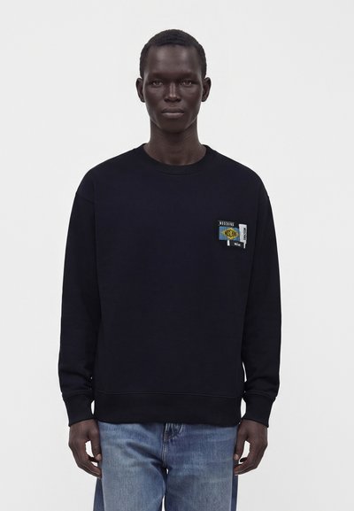 Homme portant un sweat-shirt noir avec un petit patch logo Moschino coloré sur la poitrine et un jean bleu, debout devant un fond blanc uni.