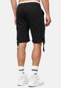 Pantalones cortos de carga negros con cintura elástica, bolsillos laterales y traseros, y cuerdas ajustables en el dobladillo. Combinados con calcetas deportivas blancas y zapatillas.
