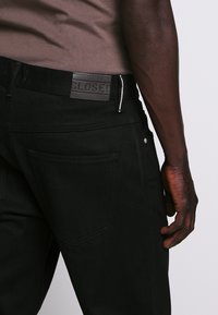 Pantalones de mezclilla negros con un acabado texturizado, que presentan una etiqueta de cuero negro, accesorios plateados y dos bolsillos traseros.