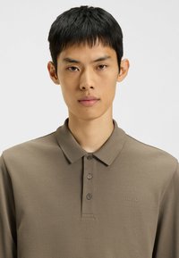 Polo marrone in tessuto strutturato con colletto, abbottonatura a tre bottoni e un logo discreto sul petto sinistro.