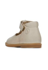 Zapato de cuero beige con punta redondeada, correa de velcro ajustable y suela de goma. Presenta detalles perforados y logo en relieve en el talón.